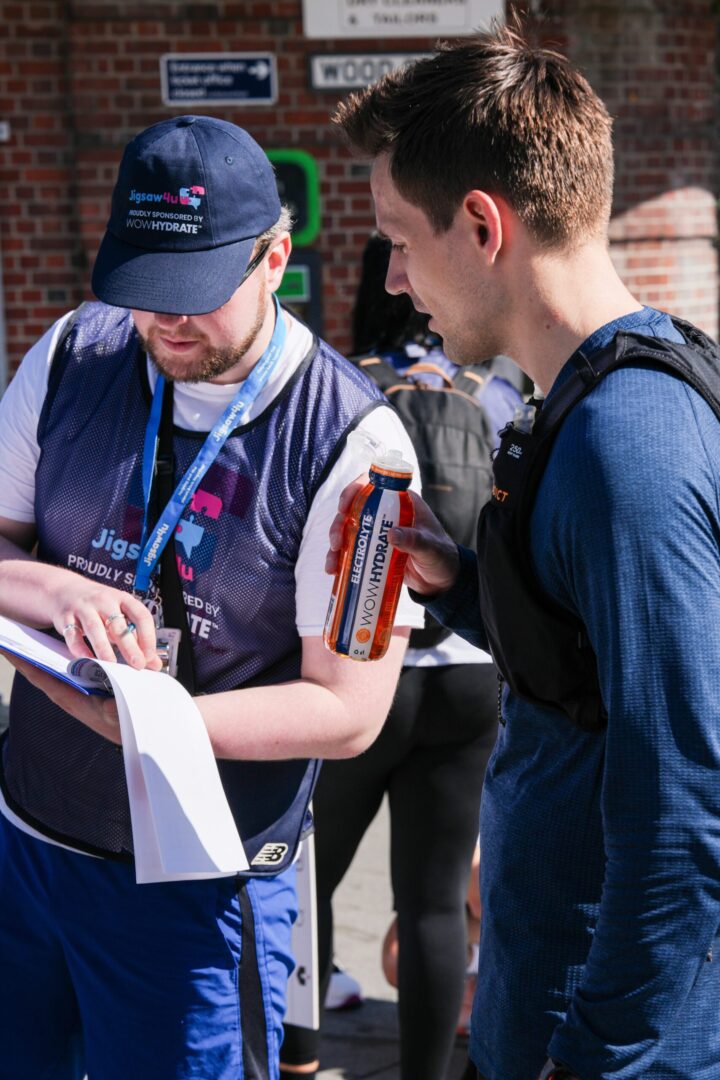 Lead walker with WOW Hydrate bib and cap on looking and sheet on a clipboard. Standing next to one of Jigsaw4u's trustees who is wearing navy